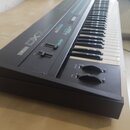 Černý digitální syntezátor s klávesnicí. Na horním panelu je bílý nápis YAMAHA DX7, několik posuvníků a dva otočné knoflíky. Povrch přístroje vykazuj…