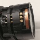Detailní pohled na vnitřek černého plastového komponentu, pravděpodobně části elektroniky nebo snímače. Je vidět měděné vinutí cívky a několik zlatis…