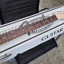 Dřevěný krk elektrické kytary s hnědým hmatníkem a světlou hlavou. Na krabici pod krkem je nápis GENUINE REPLACEMENT GUITAR NECK a logo Fender s text…