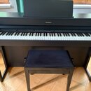 Černý digitální piano s klaviaturou a přívodní stolečkem s černým čalouněním. Na horním panelu je umístěn nápis Roland. Nástroj vykazuje čistý povrch…