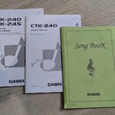Tři tištěné příručky značky CASIO na dřevěném povrchu. První bílá kniha nese nápisy CTK-240, CTK-245, USER'S GUIDE, GUÍA DEL USUARIO a v dolním rohu…