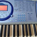 Detailní pohled na ovládací panel a klávesy elektronického klavíru. Na panelu je vidět tabulka s názvy zvuků rozdělená do kategorií jako PIANO, GUITA…