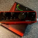 Červené audio rozhraní s nápisem Scarlett Solo. Na horním panelu jsou dva menší a jeden velký černý ovládací knoflík, tlačítka s nápisy Inst, 48V a A…