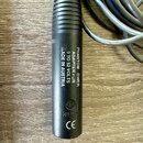Černý cylindrický adaptér s textem PHANTOM-POWER ADAPTER FOR 9 TO 52 VOLTS MADE IN AUSTRIA. Na spodní části jsou vidět symboly CE a logo s nápisem MG…