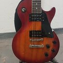 Elektrická kytara s trupem v červeně-oranžovém sunburst provedení a černým hmatníkem. Nástroj má dva černé humbuckery, černý pickguard a čtyři černé…