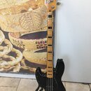 Černá elektrická basová kytara s jasným dřevěným hmatníkem a černými pražcovými značkami. Na hlavě je uveden nápis Squier a Jazz Bass. Tělo je v lesk…