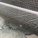 Detailní záběr na hranu černého reproduktoru nebo komba. Horní část tvoří tkaná textilní mřížka, spodní část je potažena černým texturovaným vinylem…