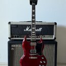 Elektrická kytara v tmavě červené barvě s černým pickguardem, umístěná na stojanu. Na hlavě hmatníku je nápis Gibson a na kombu v pozadí značka Marsh…