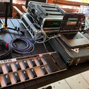 Na černém stole je rozložena sada hudebního vybavení. V popředí je stříbrný MIDI kontrolér s nápisem FCB1010 MIDI FOOT CONTROLLER a řadou černých spí…