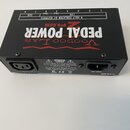 Černý napájecí adaptér pro efektové pedály s nápisy v červené a bílé barvě. Na horní straně je text Voodoo Lab PEDAL POWER 2 PLUS a schéma výstupů s…