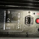 Detail zadní strany aktivního reproduktoru s černým povrchem. Na stříbrném štítku je uveden název Active Speaker-System KB-210A a technické specifika…