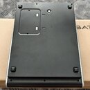 Fotografie zachycuje spodní stranu černého elektronického zařízení s kovovým rámem, které leží na hnědé kartonové krabici. Na krabici je viditelný fr…