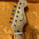 Fotografie zachycuje hlavu elektrické kytary z přírodního dřeva s viditnou sletovanou strukturou. Na hlavě je černý nápis Fender JAZZMASTER s doplňuj…