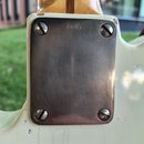 Detail zadní strany krku elektrické kytary v krémové barvě. Vidět je kovová krytka s nápisem "Luthier", připevněná čtyřmi šrouby. Lak na těle kytary…