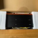 Černý elektronický přístroj s oranžovou horní hranou umístěný v přepravním kartonu s bílými polystyrenovými výplněmi. Na horní černé ploše jsou vidět…