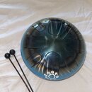 Modrý kovový steel tongue drum s vyfrézovanými jazyčky. Na spodní části nástroje je bílý nápis s označením tónů: G, G, F, #A, A, C, C, D. Vedle nástr…