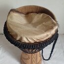 Djembe s tělem z vyřezávaného světlého dřeva a membránou z přírodní kůže s viditelným chlupem po okrajích. Membrána je upevněna černým pleteným prová…