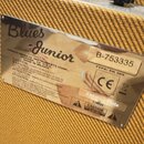 Detail zadní strany kytarového komba potaženého žlutým texturovaným materiálem. Na kovovém štítku je nápis Blues Junior, A PRODUCT OF FENDER MUSICAL…