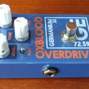 Modrý kytarový efekt pedál s nápisy OXBLOOD, OVERDRIVE a GERMANIUM 32. Na horní straně jsou tři bílé oválné ovladače s popisky DRIVE, TONE a LEVEL, j…