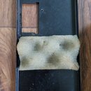 Černý plastový nebo kovový kryt s obdélníkovým výřezem v horní části. Uprostřed je přilepen kus šedé, mírně znečištěné houbové pěny. Povrch krytu vyk…