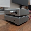 Dvě stohované komponenty v šedých kovových šasi s matným povrchem. Horní jednotka má stříbrný ovládací knoflík, tlačítko napájení a konektor pro sluc…
