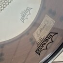 Detailní pohled na bílý bubenový kožen s černým lemováním. Na povrchu je černý nápis REMO v rámci koruny a text AMBASSADOR SNARE SIDE MADE IN U.S.A.…