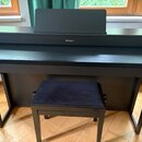 Černý digitální piano v dřevěném korpusu s přiloženým černým polstrovaným stoličkou. Na přední hraně nad klávesnicí je bílý nápis Roland. Nástroj má…