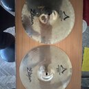 Dvě mosazné činely položené na hnědé dřevěné desce. Oba mají nápisy A Custom, MASTERROUND HI HAT a Avid Zildjian. Povrch činelů je klepaný, horní kus…