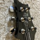Detail hlavy černé elektrické kytary s chromovanými ladičkami. Na povrchu je bílý nápis „Paul Reed Smith SE“ a v dolní části nápis „Custom“. Lak je g…