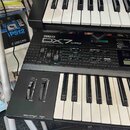Černý digitální syntetizátor s bílými a černými klávesami. Na horním panelu je bílý nápis YAMAHA a model DX7 IIFD s doplňkem DIGITAL PROGRAMMABLE POL…