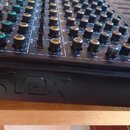 Detailní pohled na černý mixážní pult s řadou ovládacích knoflíků v různých barvách (černá, zelená, žlutá). Na přední hraně šasi je vyražen nápis "mo…