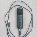 Černý kondenzátorový mikrofon s mřížkou a integrovaným USB kabelem. Na těle jsou nápisy MUTE, GAIN, HEADPHONES a označení modelu KM-2U. Ve spodní čás…