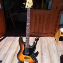 Elektrická basová kytara s trupem v přechodové barvě od černé přes červenou k žluté (sunburst) a černým pickguardem. Na hlavě hmatníku je vidět logo…