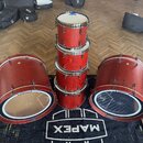 Sada červených bubnů s kovovým hardwarem umístěná na černém koberci s bílým nápisem MAPEX. Sestává ze dvou velkých basových bubnů, jednoho tom-tomu s…