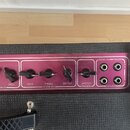 Detail ovládacího panelu kytarového komba v růžové barvě s černým texturovaným potahem. Panel obsahuje pět černých oválných knoflíků s popisky LEVEL,…