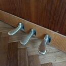 Detailní pohled na tři mosazné pedály klaviru. Pedály jsou umístěny v dřevěné liště světlé barvy, nad kterou je vidět tmavší dřevěný povrch nástroje.…