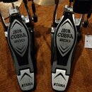 Dva bicí pedály značky TAMA z řady IRON COBRA 200. Pedály mají kovové stříbrné tělo s černými vložkami a bílými nápisy IRON COBRA 200 a TAMA. Povrch…