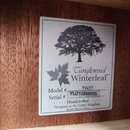 Detailní záběr na bílý štítek s černým tiskem, který je nalepený na dřevěném povrchu. Štítek obsahuje siluetu stromu a nápisy Tanglewood Winterleaf™,…