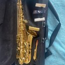Zlatý saxofon v černém otevřeném pouzdře. K nástroji je přiložen mouthpiece, dvě krabičky rákosů značky Rico a bílý předmět v plastovém obalu. Na pou…