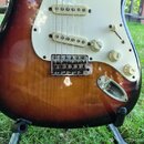 Detail elektrické kytary s tělem v barevném přechodu od černé přes hnědou po žlutou. Nástroj má bílý pickguard, tři snímače s bílými krytkami a tři b…