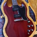 Elektrokytara s tmavě červeným lakovaným povrchem a černým pickguardem v hnědém plišovém kufru. Nástroj má dva humbuckery, stříbrný most a čtyři ovlá…
