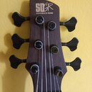 Hlava basové kytary z tmavého dřeva s šesti ladicími kolíky. V horní části je bílý nápis SDGR a pod ním text SOUNDGEAR BY IBANEZ. Povrch dřeva vykazu…