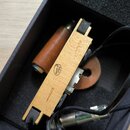 V černé krabici s pěnovou výplní je umístěn dřevěný kousek příslušenství s nápisem MADE IN CHINA v bílém kruhu. Vedle něj se nachází hnědý kožený vál…