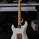 Zadní strana bílé elektrické kytary s hnedým javorovým hmatníkem a hlavou. Na zadní části krku je vidět logo "Fender" v kovovém rámečku. Tělo kytary…