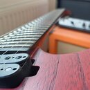 Detailní pohled na hmatník a tělo elektrické kytary v červené barvě s viditnou strukturou dřeva. Na černém snímači je částečně viditelný nápis "MANSO…
