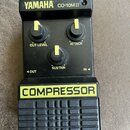 Černý kompresor s nápisem YAMAHA CO-10M.II v horní části a žlutým nápisem COMPRESSOR na spodním panelu. Zařízení má tři černé oválné knoflíky s žlutý…