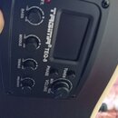 Detailní pohled na ovládací panel akustické kytary s černým plastovým povrchem. Panel obsahuje čtyři otočné knofíky pro ekvalizaci s nápisy BASS, MID…
