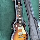 Elektrická kytara typu Les Paul s barevným přechodem od žluté k tmavě hnědé v černém tvrdém kufru s šedým polstrováním. Na hlavě hmatníku je bílý náp…