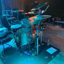 Celá bicí souprava s několika cymbály, tommy a basovým bubnem v kovovém povrchu. Nástroje jsou umístěny na černém koberci vedle černé židličky a otev…
