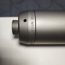Detailní pohled na horní část stříbřitého kondenzátorového mikrofonu s kovovým povrchem. Na těle je vidět nápis „MADE NT1-A“ a pod ním začátek označe…